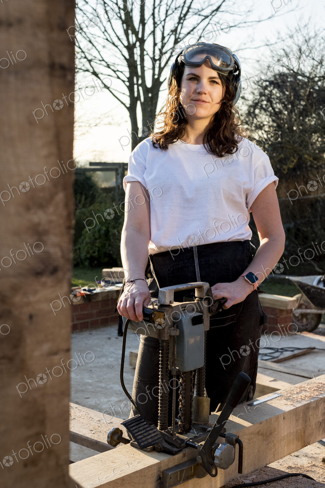 Woman wearing protective goggles holding machine, working on wooden building frame.