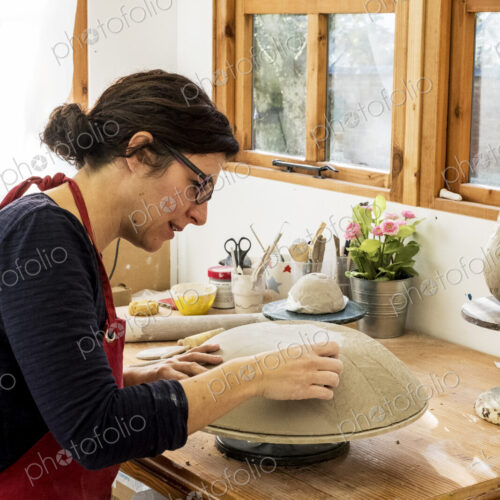 Woman wearing red apron sitting in her ceramics workshop, working on clay bowl.