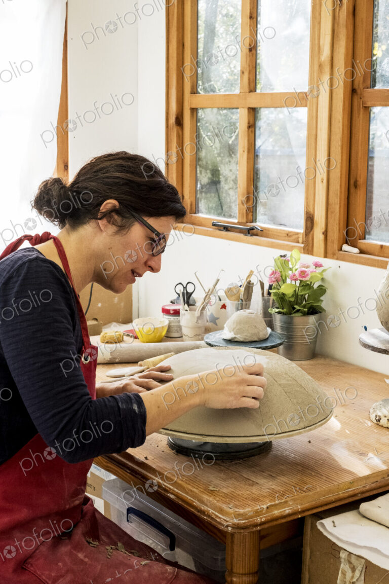 Woman wearing red apron sitting in her ceramics workshop, working on clay bowl.