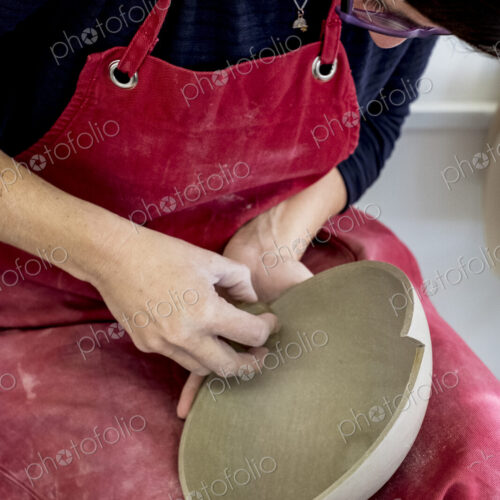 Woman wearing red apron sitting in her ceramics workshop, working on clay bowl.
