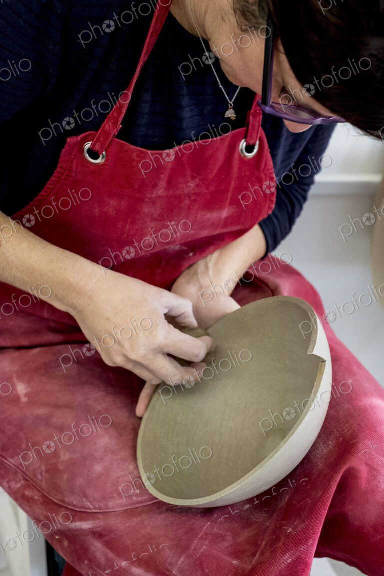 Woman wearing red apron sitting in her ceramics workshop, working on clay bowl.