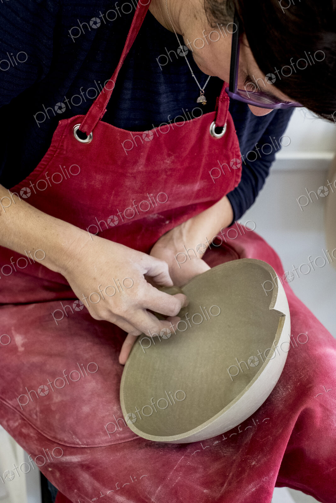 Woman wearing red apron sitting in her ceramics workshop, working on clay bowl.