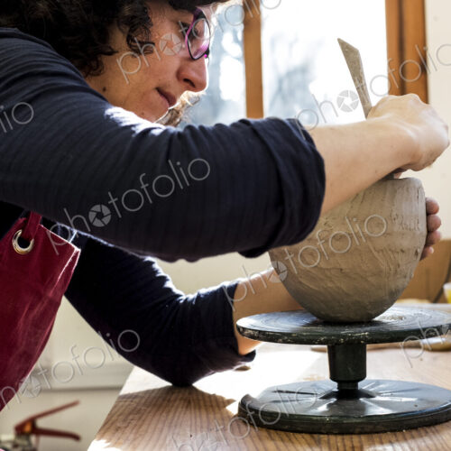 Woman wearing red apron sitting in her ceramics workshop, working on clay vase.