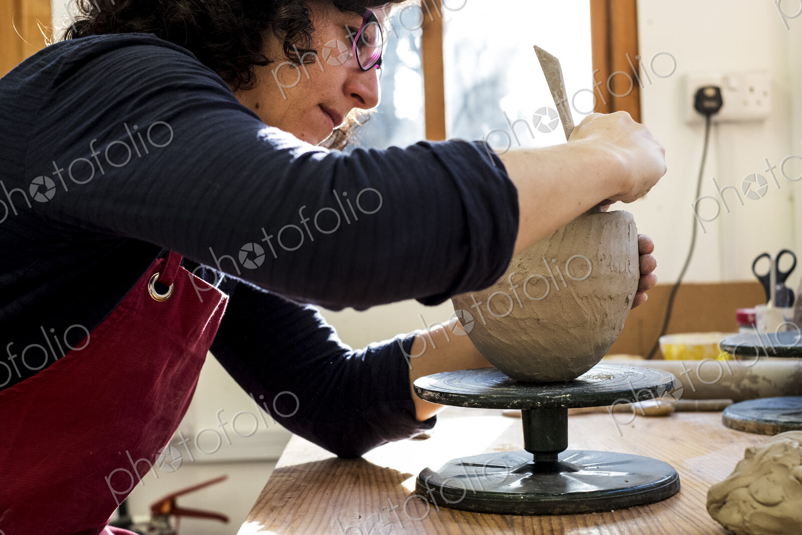 Woman wearing red apron sitting in her ceramics workshop, working on clay vase.