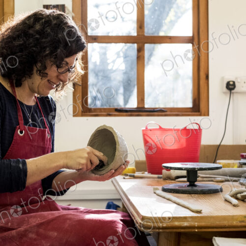 Woman wearing red apron sitting in her ceramics workshop, working on small clay bowl.