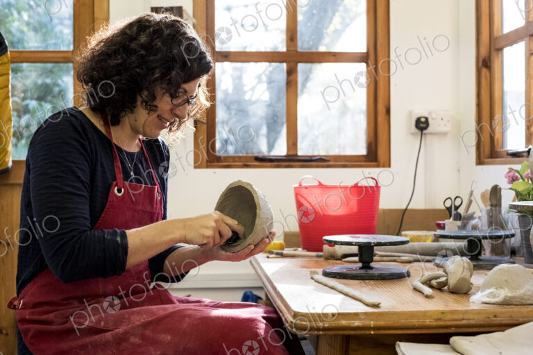 Woman wearing red apron sitting in her ceramics workshop, working on small clay bowl.