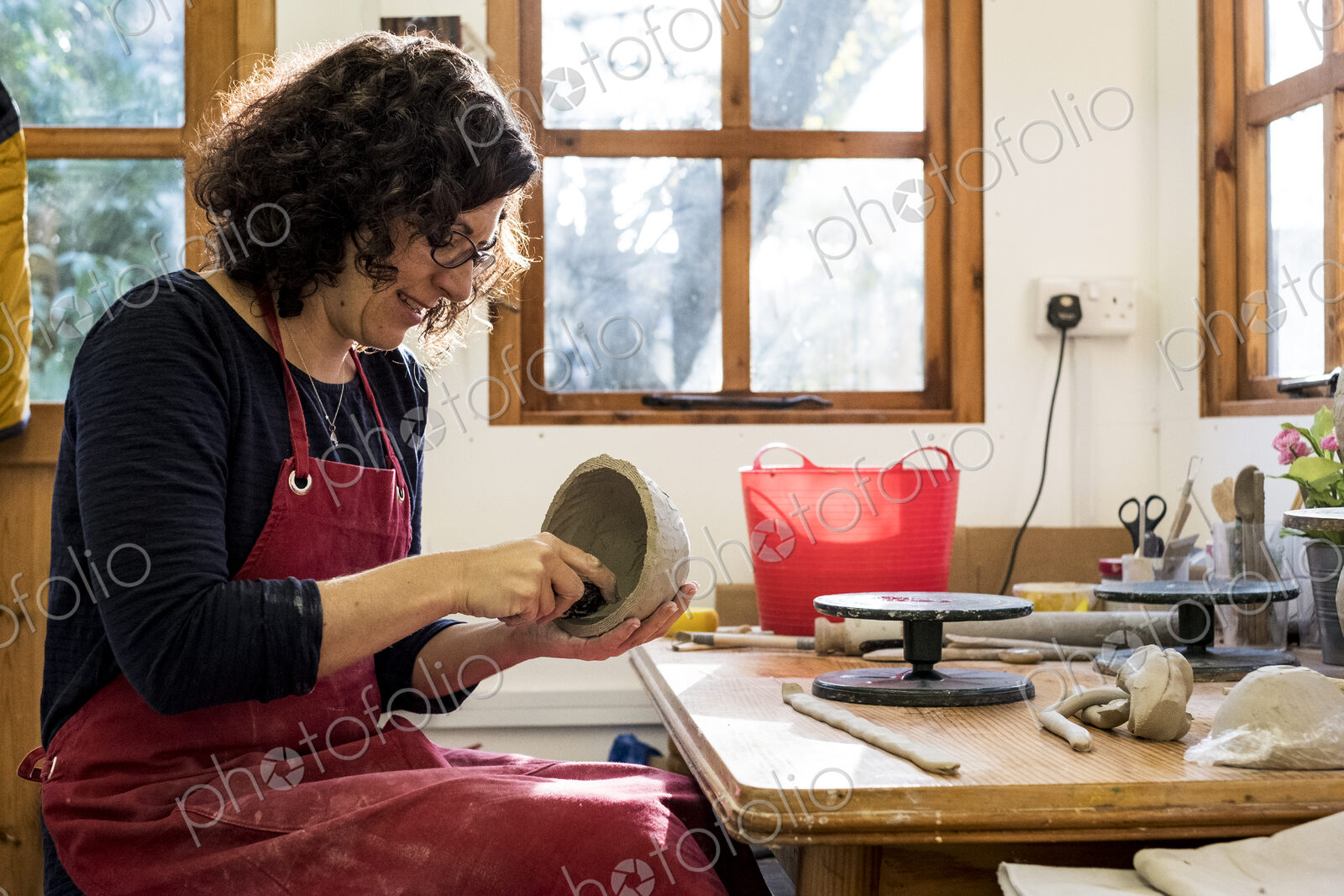 Woman wearing red apron sitting in her ceramics workshop, working on small clay bowl.