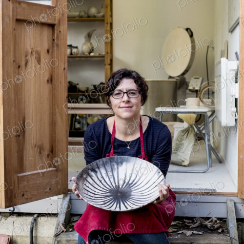 Woman wearing red apron sitting on steps outside her workshop, holding ceramic bowl…