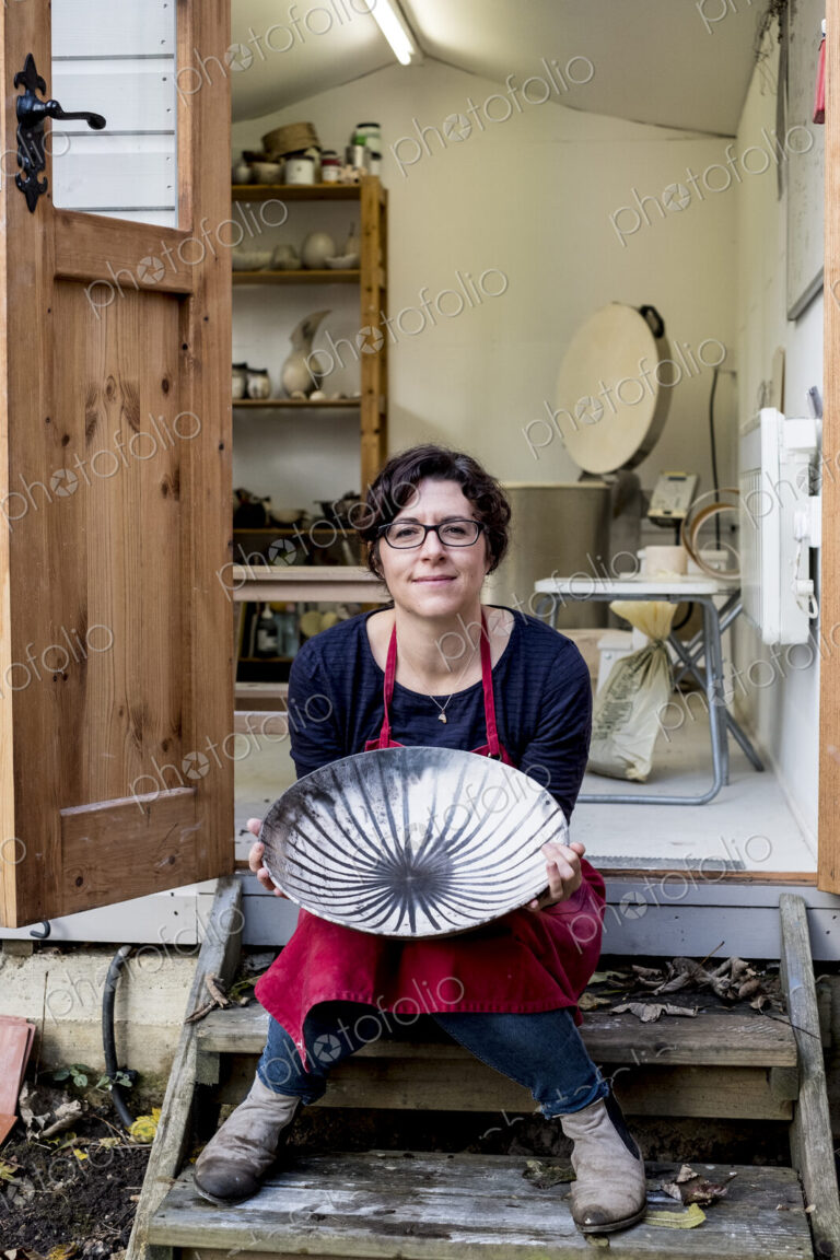 Woman wearing red apron sitting on steps outside her workshop, holding ceramic bowl…