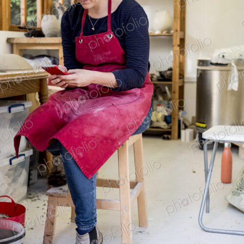 Woman wearing red apron sitting on stool in her ceramics workshop, checking her mobile…