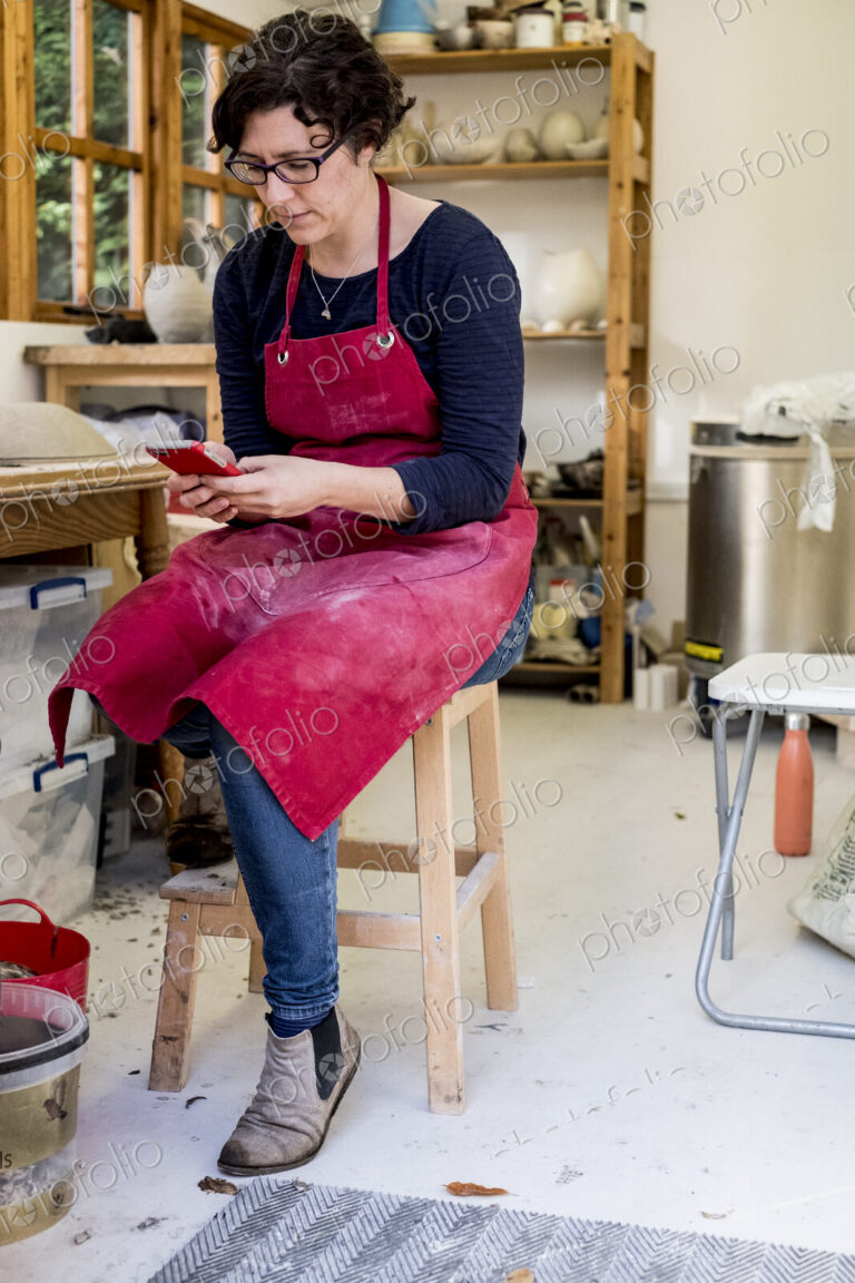 Woman wearing red apron sitting on stool in her ceramics workshop, checking her mobile…