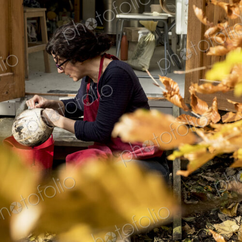 Woman wearing red apron sitting outside her ceramics workshop, holding vase.