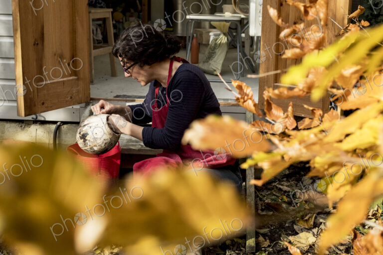 Woman wearing red apron sitting outside her ceramics workshop, holding vase.