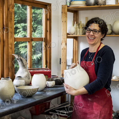 Woman wearing red apron standing in her workshop, holding ceramic vase, smiling at camera.