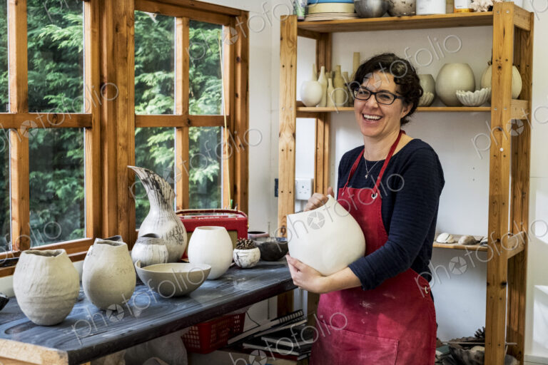 Woman wearing red apron standing in her workshop, holding ceramic vase, smiling at camera.