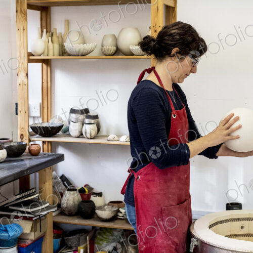 Woman wearing red apron standing in her workshop next to kiln, holding ceramic vase.