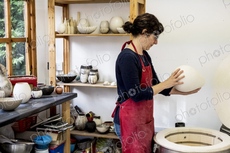 Woman wearing red apron standing in her workshop next to kiln, holding ceramic vase.