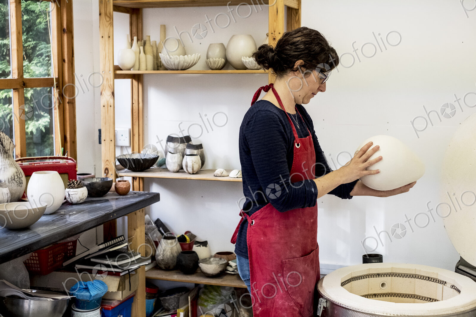 Woman wearing red apron standing in her workshop next to kiln, holding ceramic vase.