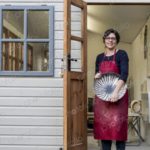 Woman wearing red apron standing on steps outside her workshop, holding ceramic bowl…