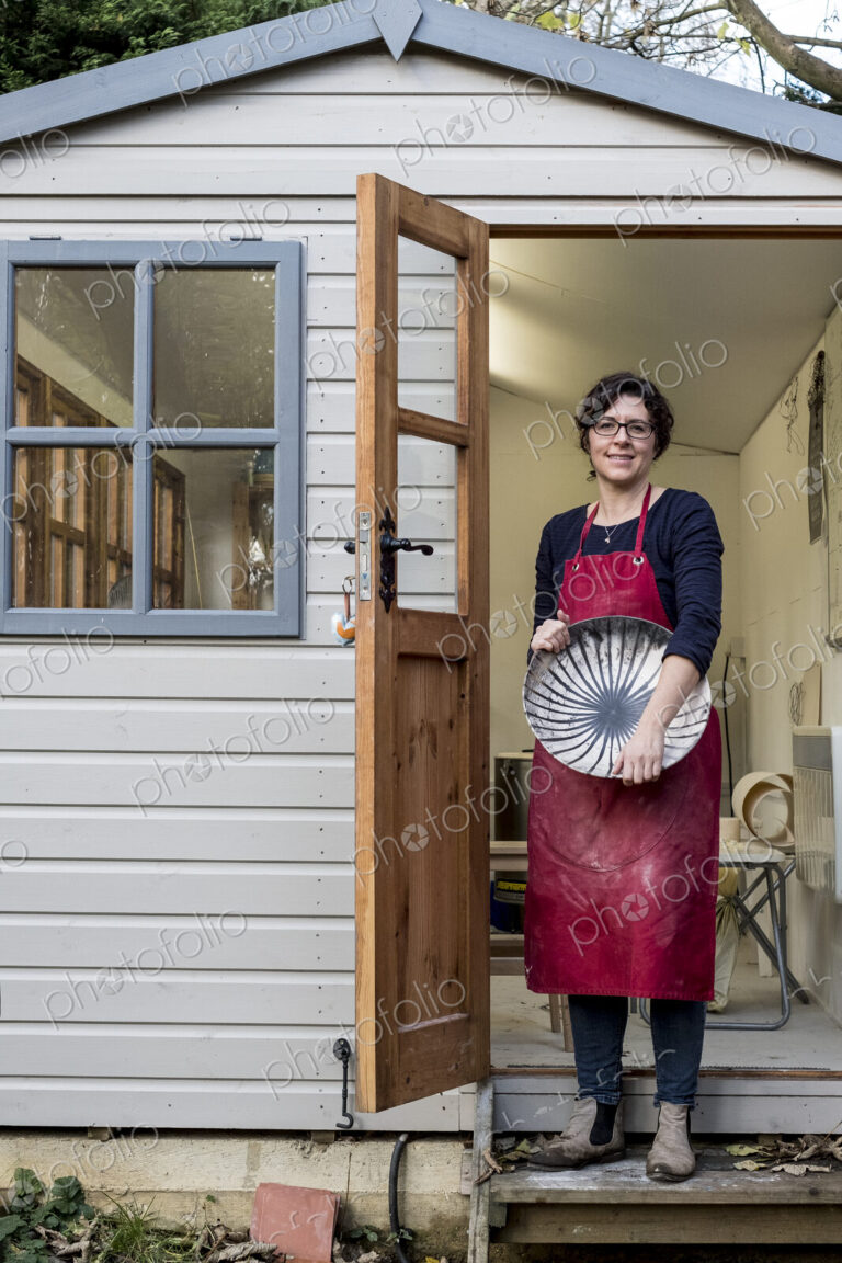 Woman wearing red apron standing on steps outside her workshop, holding ceramic bowl…