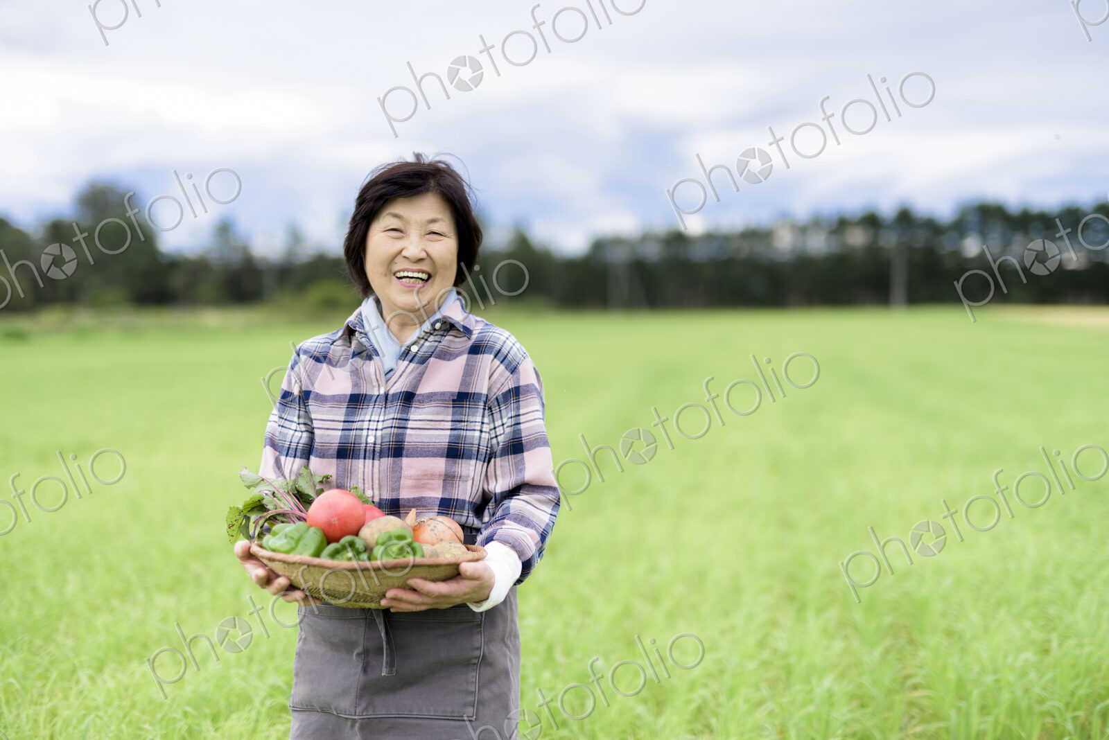 Woman with black hair wearing checkered shirt standing in a field, holding basket with…