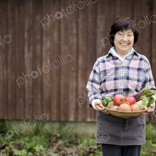 Woman with black hair wearing checkered shirt standing in a garden, holding basket… Woman with black hair wearing checkered shirt standing in a garden, holding basket…