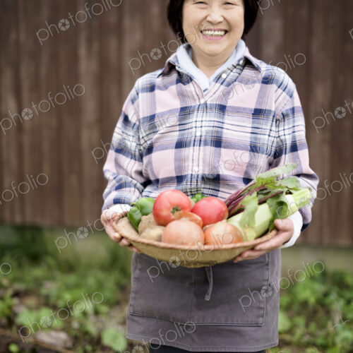 Woman with black hair wearing checkered shirt standing in a garden, holding basket… Woman with black hair wearing checkered shirt standing in a garden, holding basket…