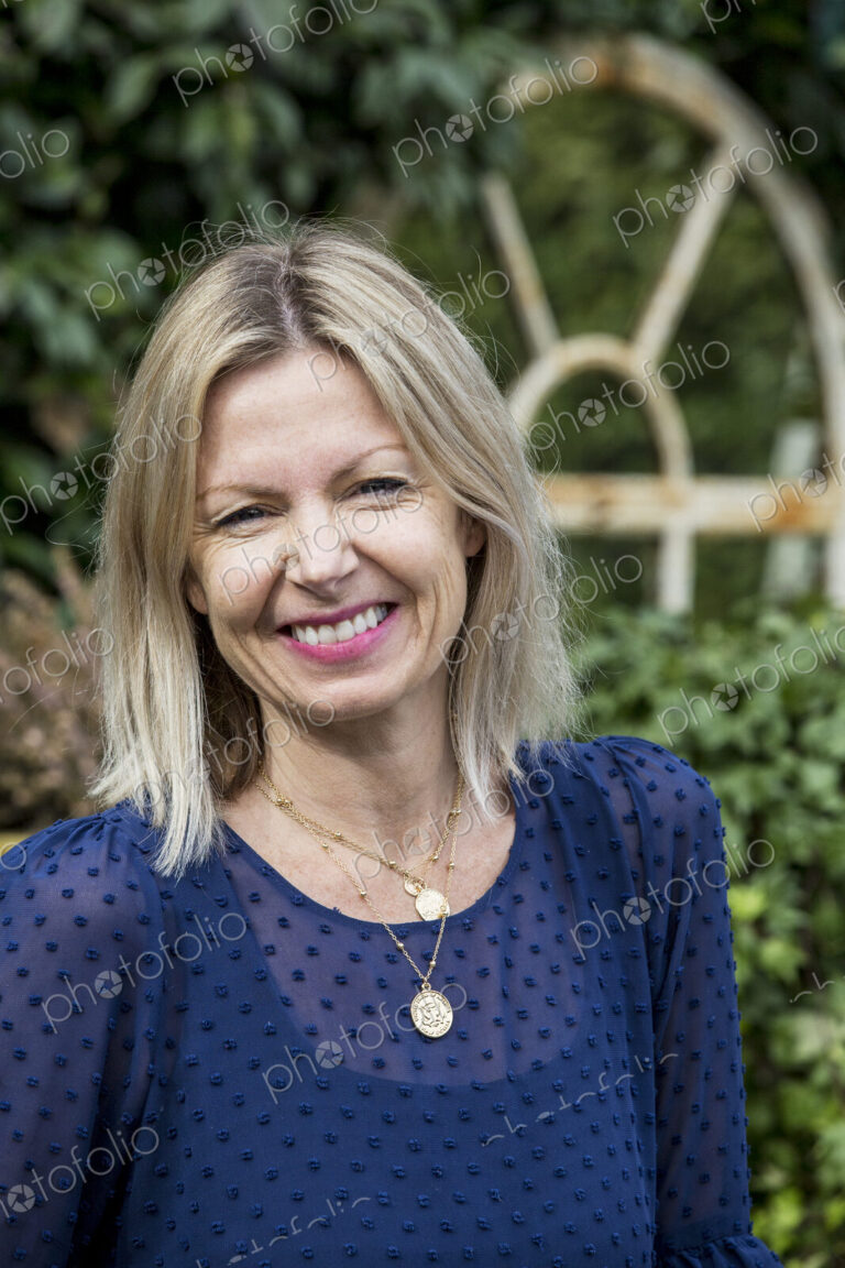 Woman with blond hair and wearing blue top standing outdoors, smiling at camera.