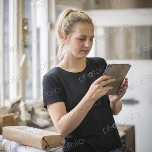 Woman with blond hair standing in a workshop using a digital tablet, wrapped parcels… Woman with blond hair standing in a workshop using a digital tablet, wrapped parcels…