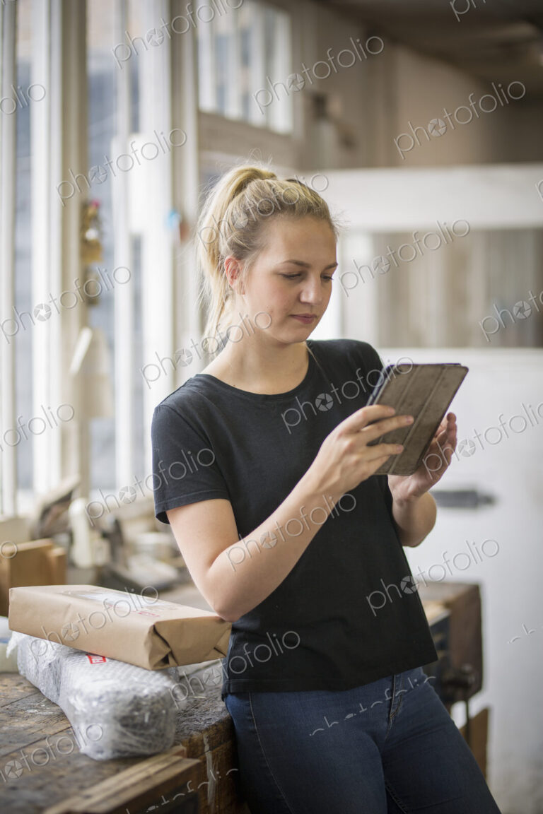 Woman with blond hair standing in a workshop using a digital tablet, wrapped parcels…