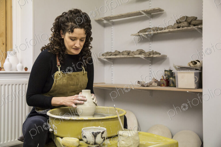 Woman with brown curly hair wearing apron shaping clay vase on pottery wheel.