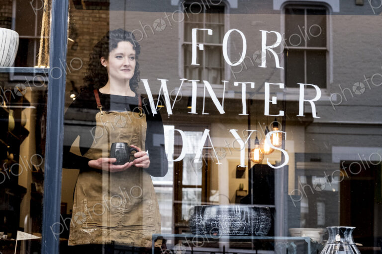 Woman with brown curly hair wearing apron standing behind window of pottery shop,…