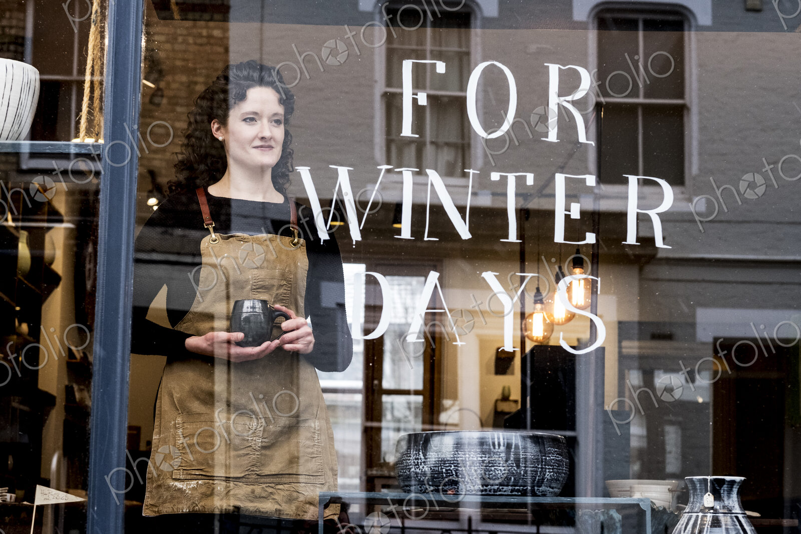 Woman with brown curly hair wearing apron standing behind window of pottery shop,…
