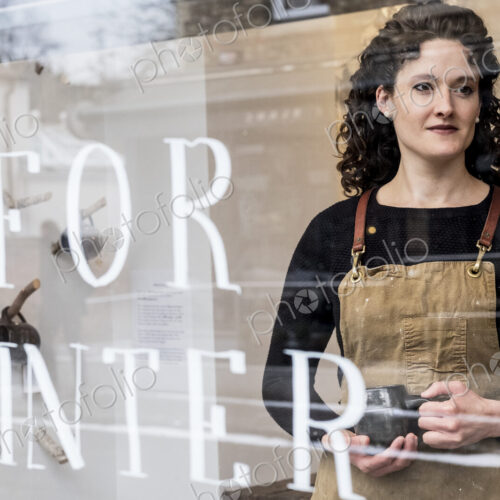 Woman with brown curly hair wearing apron standing behind window of pottery shop,… Woman with brown curly hair wearing apron standing behind window of pottery shop,…