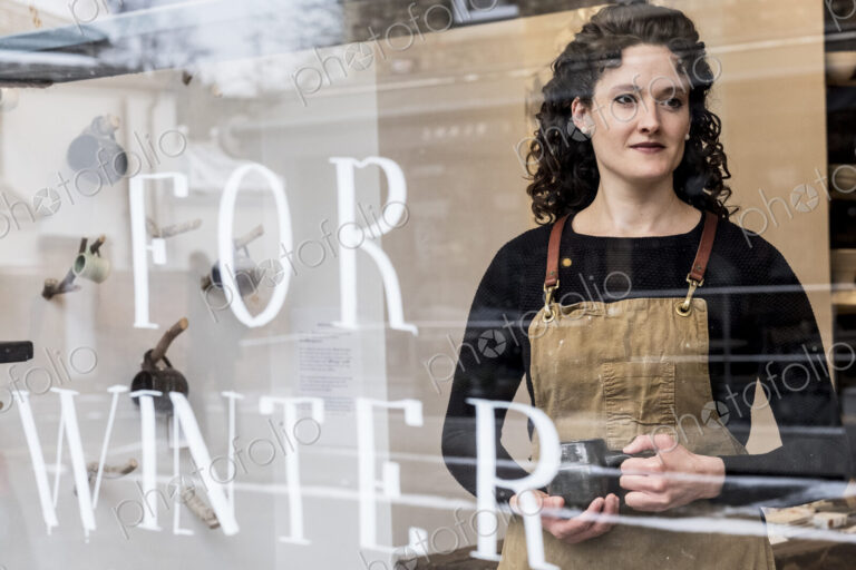 Woman with brown curly hair wearing apron standing behind window of pottery shop,…
