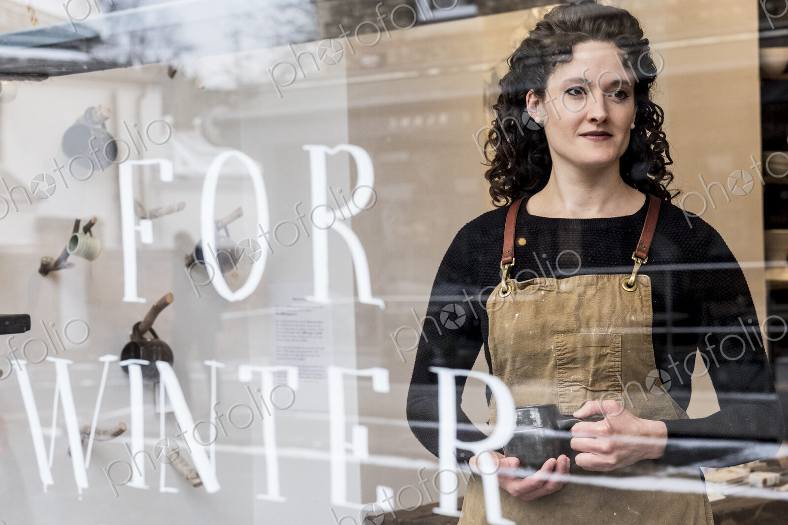 Woman with brown curly hair wearing apron standing behind window of pottery shop,…