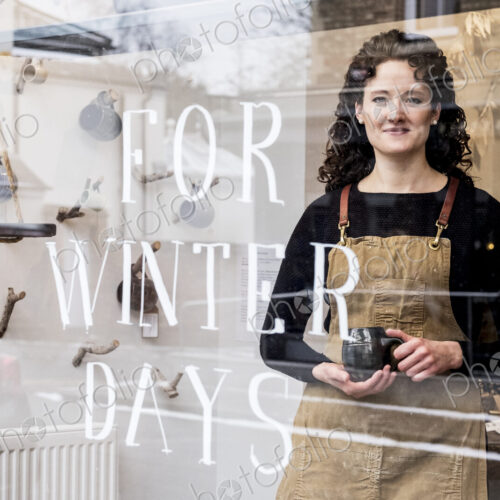 Woman with brown curly hair wearing apron standing behind window of pottery shop,… Woman with brown curly hair wearing apron standing behind window of pottery shop,…