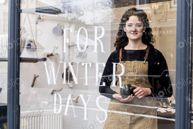 Woman with brown curly hair wearing apron standing behind window of pottery shop,…