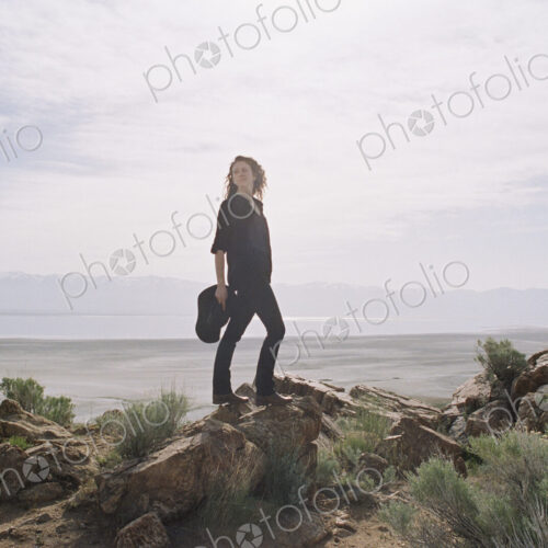 Woman with brown, long and curly hair standing on rocks, a lake and mountains in the… Woman with brown, long and curly hair standing on rocks, a lake and mountains in the…