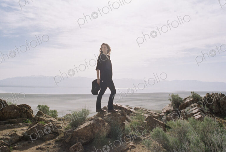 Woman with brown, long and curly hair standing on rocks, a lake and mountains in the…