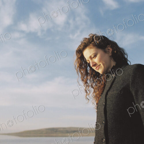 Woman with brown, long and curly hair standing on the shore of a lake.
