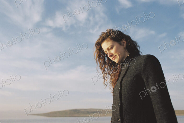 Woman with brown, long and curly hair standing on the shore of a lake.