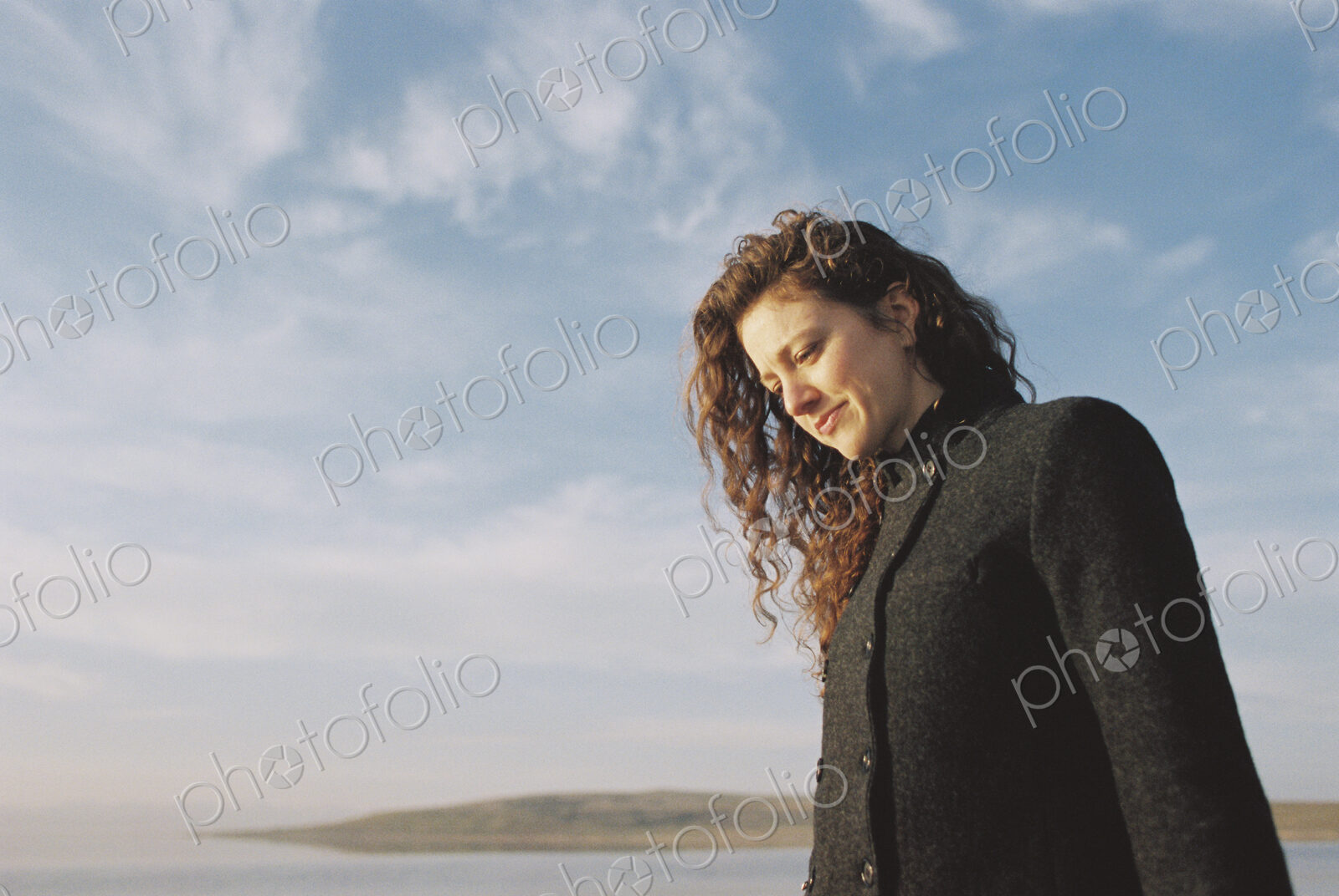 Woman with brown, long and curly hair standing on the shore of a lake.