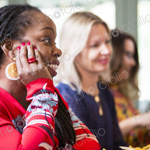 Woman with cornrows wearing golden earrings and red top sitting at a table, listening… Woman with cornrows wearing golden earrings and red top sitting at a table, listening…