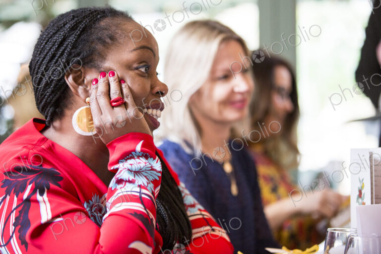 Woman with cornrows wearing golden earrings and red top sitting at a table, listening…