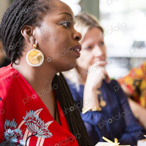 Woman with cornrows wearing golden earrings and red top sitting at a table, listening… Woman with cornrows wearing golden earrings and red top sitting at a table, listening…