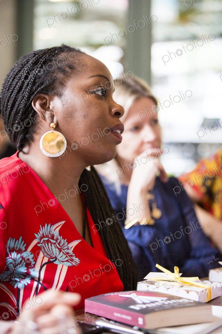 Woman with cornrows wearing golden earrings and red top sitting at a table, listening…