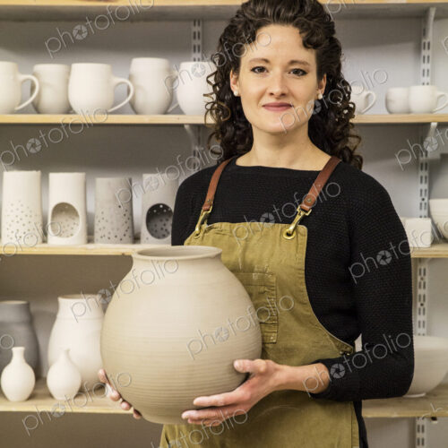 Woman with curly brown hair wearing apron holding unfired spherical clay vase. Woman with curly brown hair wearing apron holding unfired spherical clay vase.