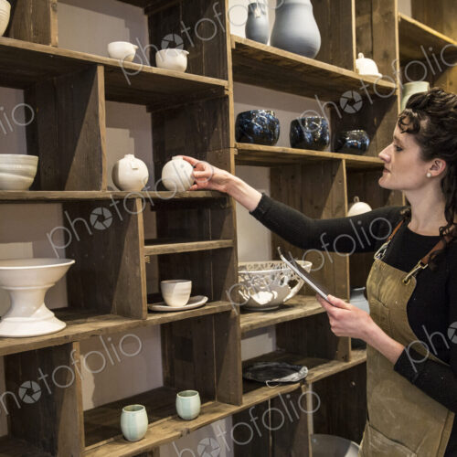 Woman with curly brown hair wearing apron standing in her pottery shop, arranging… Woman with curly brown hair wearing apron standing in her pottery shop, arranging…