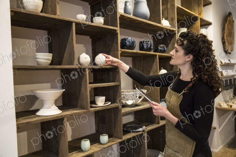 Woman with curly brown hair wearing apron standing in her pottery shop, arranging…